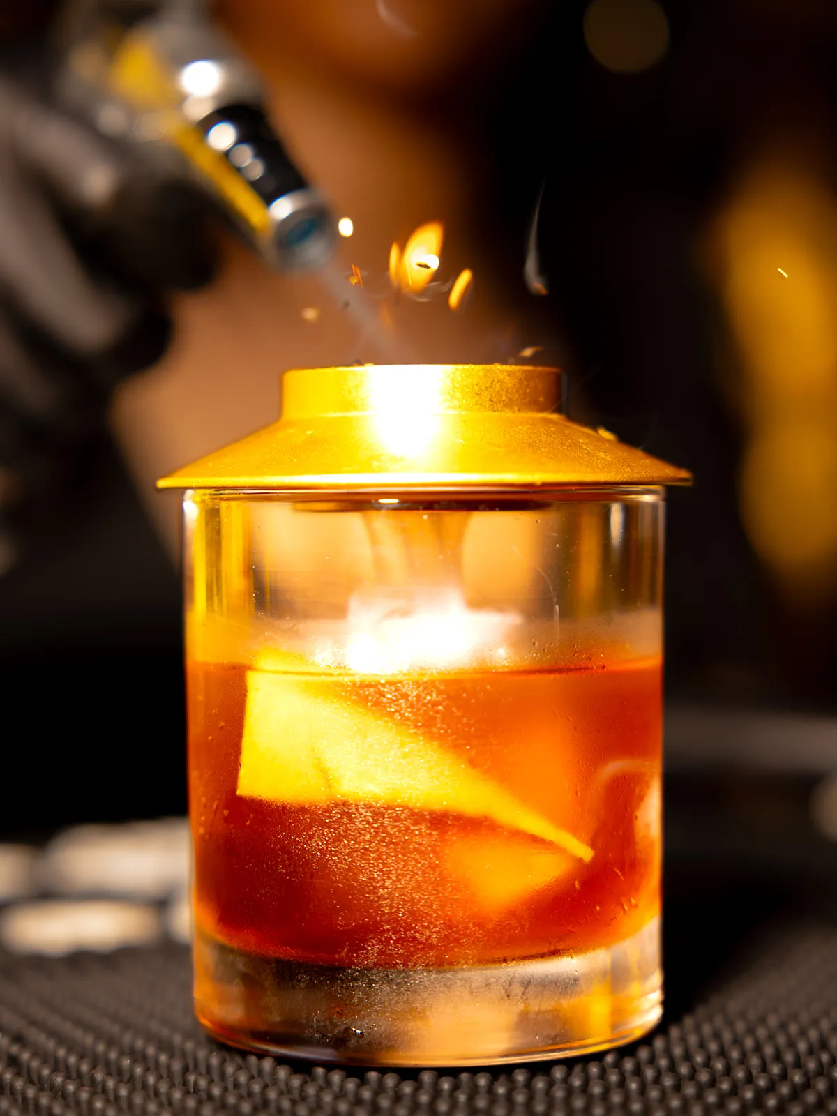 Smoked old fashioned under a brass cloche · smoke rising through the lid · torch flame above · Allure